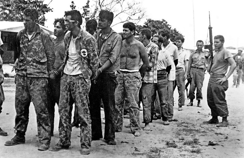 Captured members of Brigade 2506, some in camouflage, standing in a line on sandy ground, watched by an armed Cuban soldier.