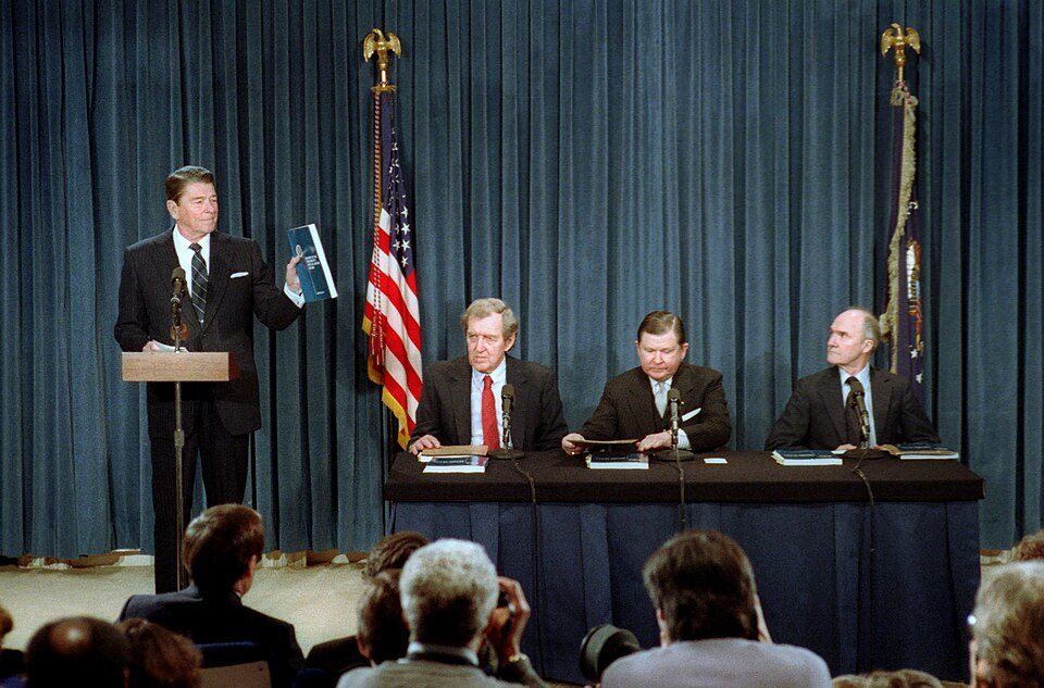 President Reagan at a podium beside John Tower, Edmund Muskie and Brent Scowcroft as he receives their report.