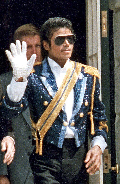 Michael Jackson in 1984, waving with a white sequined glove, wearing a sequined jacket with a gold sash and aviator sunglasses.