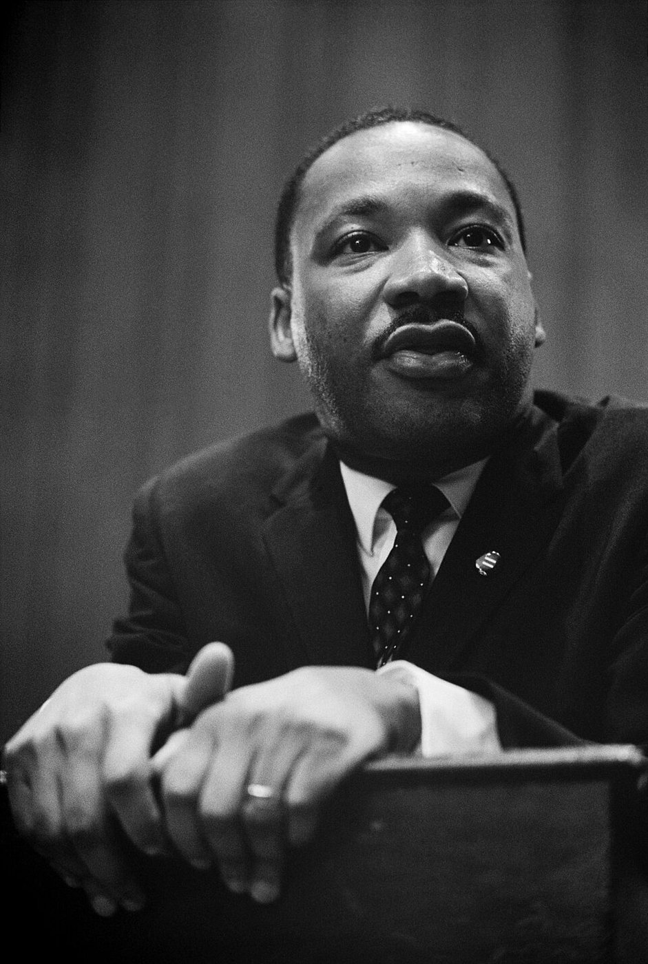 Dr. Martin Luther King Jr. at a press conference, leaning forward on a podium, in a dark suit and tie.