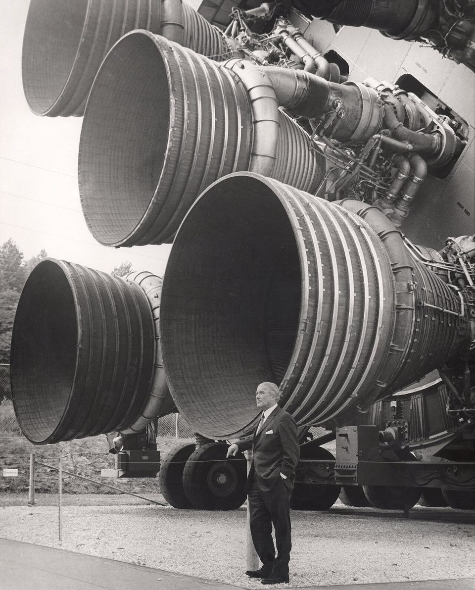 Wernher von Braun standing in front of the five huge engines at the base of a Saturn V rocket stage.