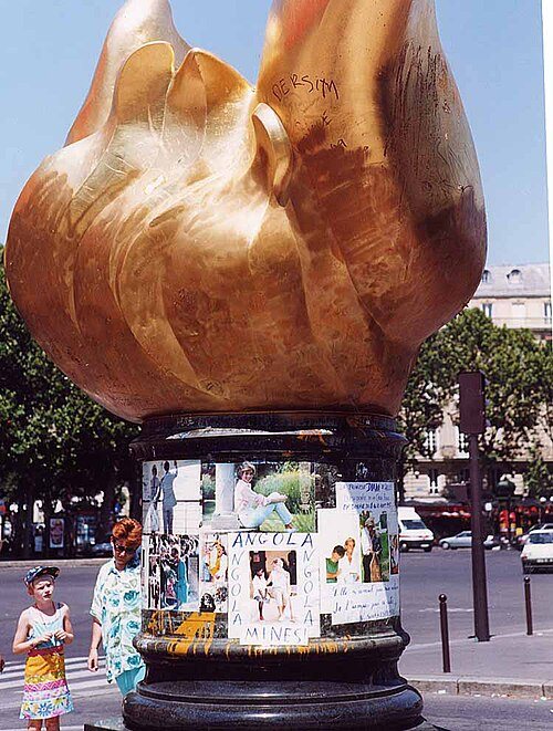 A large gilded flame sculpture on a pedestal covered with photos and notes left for Diana, with visitors standing below.