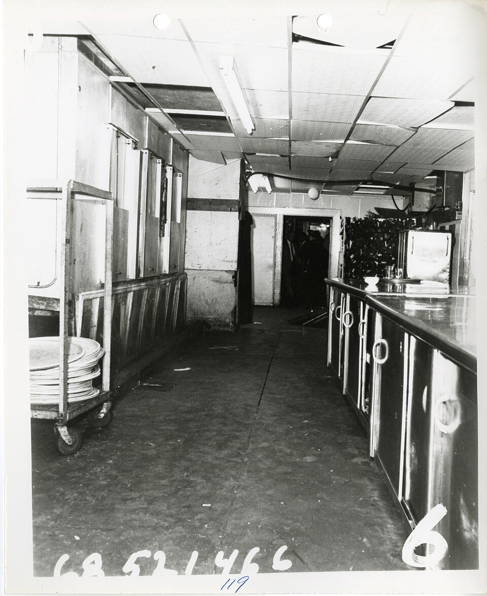 A black-and-white police photo of the Ambassador Hotel's narrow kitchen pantry: a long steel counter, a cart of plates and a low paneled ceiling, with an evidence number written along the bottom.