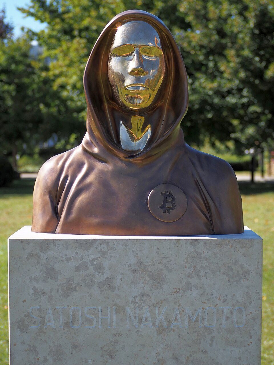 A bronze bust with a hooded figure and a smooth, reflective face, standing in a park in Budapest.