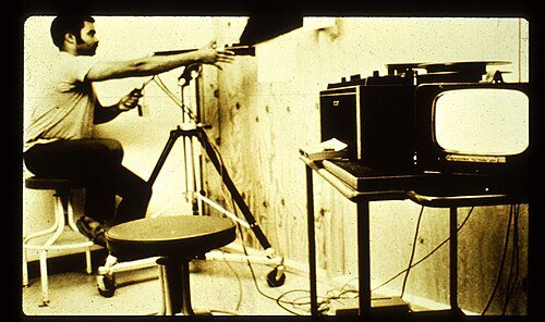 A researcher operating a film camera on a tripod beside a television monitor and recording equipment, 1971.