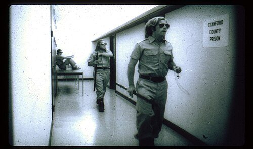 Guards in khaki uniforms and sunglasses in a basement hallway, under a sign reading Stanford County Prison.
