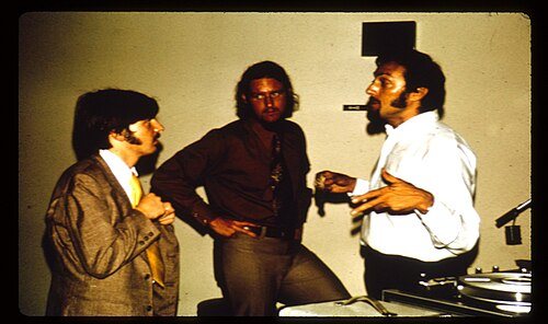 Philip Zimbardo, bearded and gesturing, talks with two young researchers next to recording equipment in 1971.